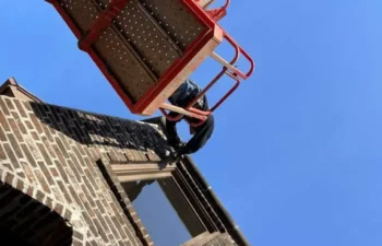 A worker in a boom lift repairs the upper section of a brick building against a clear blue sky.