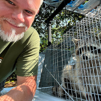 A person with a gray beard smiling next to a raccoon inside a metal cage outdoors.