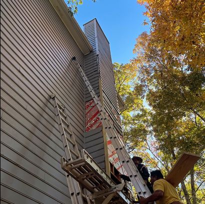 Two men work on a ladder beside the exterior of a multi-story house surrounded by trees with fall foliage.