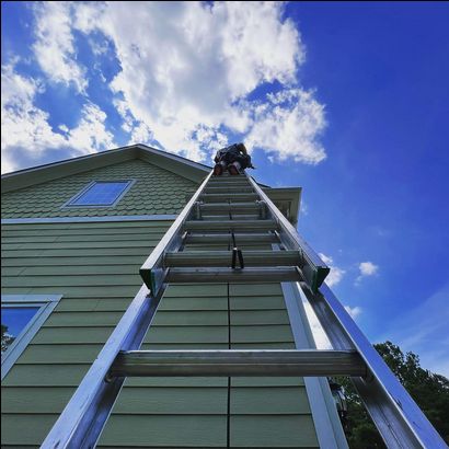 A long ladder leaning against a house with a green exterior. A person is at the top of the ladder under a partly cloudy blue sky.