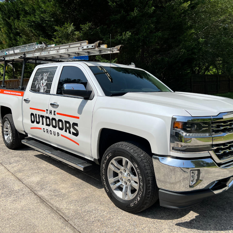 A white truck with "The Outdoors Group" logo on its side is parked on a driveway. The truck has multiple ladders secured on its rack. Trees and a wooden fence are in the background.