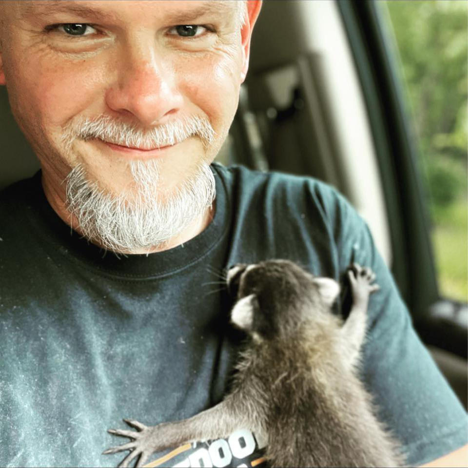 A man with a short white beard smiles while holding a small raccoon on his chest inside what appears to be a vehicle.